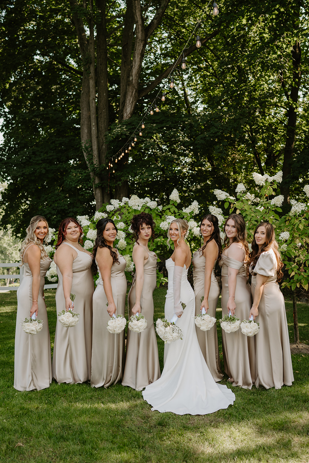 Bride and bridesmaids in champagne satin gowns smile outside under trees, holding white rose bouquets.